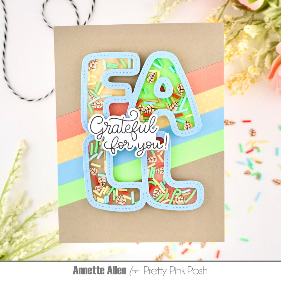 A handmade greeting card featuring layered die-cut letters spelling 'FALL' filled with a colorful autumn leaf pattern, layered over diagonal pastel stripes and a central script reading 'Grateful for you!'. The card is photographed on a white surface with decorative dried flowers and sprinkles.