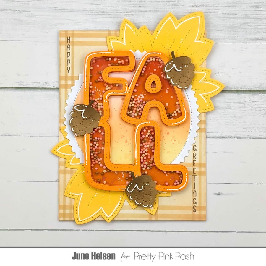 An autumn-themed greeting card featuring the word 'FALL' in large orange letters filled with tiny white dots, layered over yellow leaf shapes and brown acorns on a plaid cardstock background. The card is photographed against a light gray wood-plank backdrop, with 'June Helsen for Pretty Pink Posh' printed below.