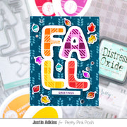 A handmade greeting card featuring the word 'FALL' in large, colorful plaid letters outlined in white, surrounded by small autumn leaf stickers on a dark teal background. The card is displayed in a styled flat-lay arrangement with craft tools and ink pads in the background.