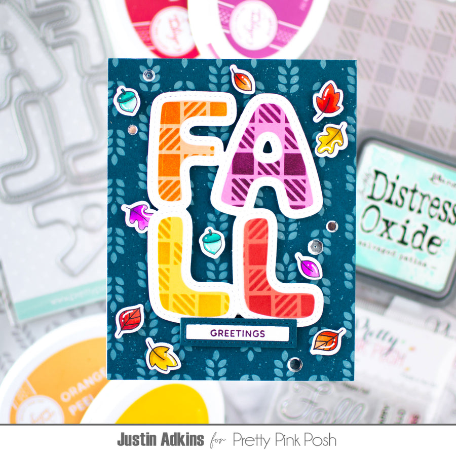 A handmade greeting card featuring the word 'FALL' in large, colorful plaid letters outlined in white, surrounded by small autumn leaf stickers on a dark teal background. The card is displayed in a styled flat-lay arrangement with craft tools and ink pads in the background.