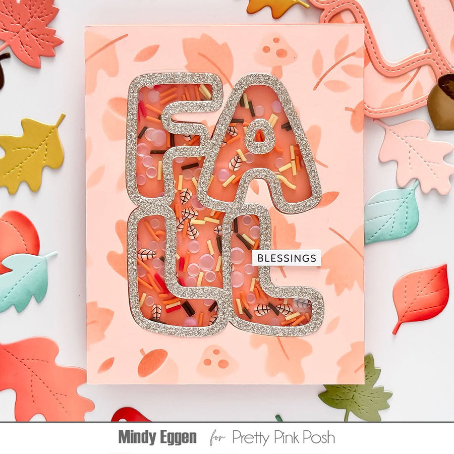 A handmade greeting card featuring the word 'FALL' in glittery silver letters with a shaker window filled with autumn-themed confetti, leaves, and acorns. The card is pink with a subtle leaf pattern, surrounded by scattered paper autumn leaves.