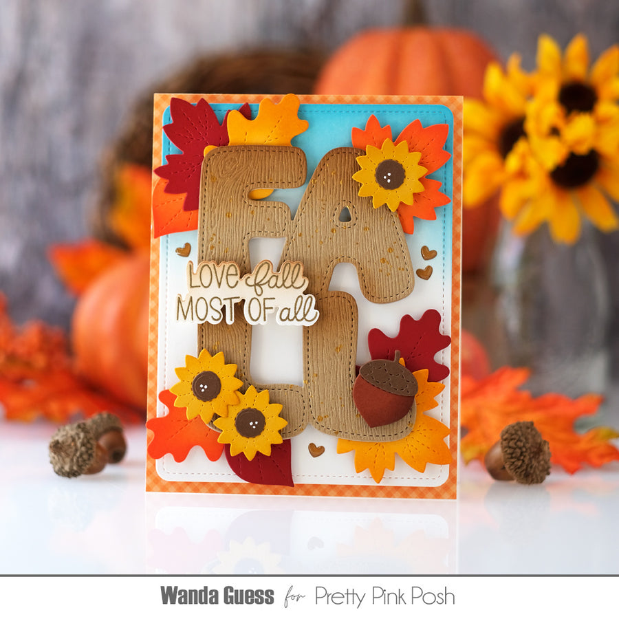 A handmade autumn-themed greeting card featuring a large layered 'FALL' design with sunflowers, fall leaves, acorns, and the phrase 'Love Fall MOST OF all'. The card is styled on a reflective white surface with pumpkins, sunflowers, and acorns in the background.