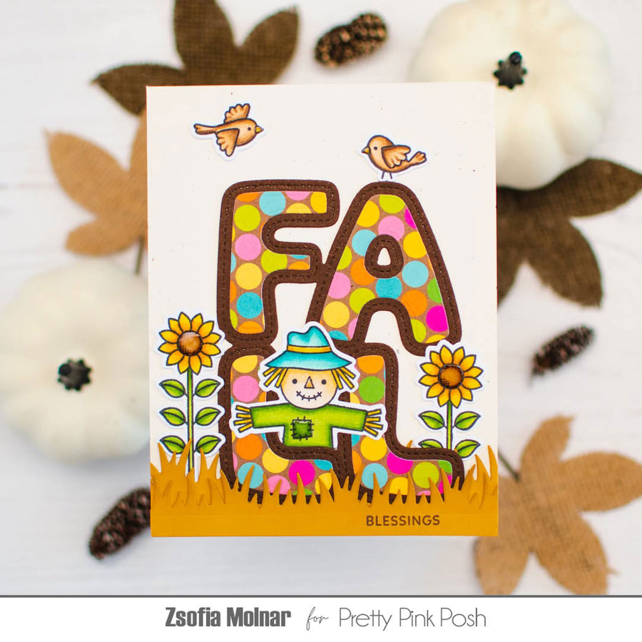 A handmade autumn-themed greeting card featuring the word 'FALL' in colorful polka-dot letters with a scarecrow, sunflowers, and birds, styled with pumpkins and fall leaves on a white surface.