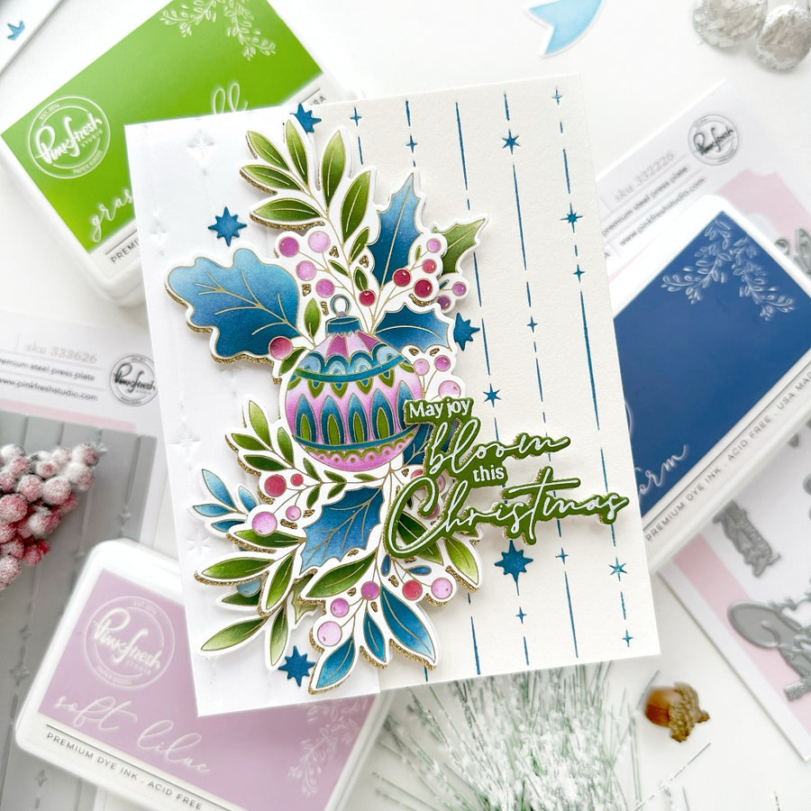 A handmade Christmas greeting card featuring a colorful stamped ornament, holly leaves, pinecones, and the phrase 'May my blossom this Christmas' in green script, styled on a white surface with crafting supplies.