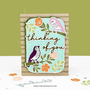 A handmade greeting card featuring two stylized birds perched on branches with the phrase 'thinking of you' in script. The card has a light aqua center panel layered over a tan striped backing, with orange and pink flowers and green leaves.