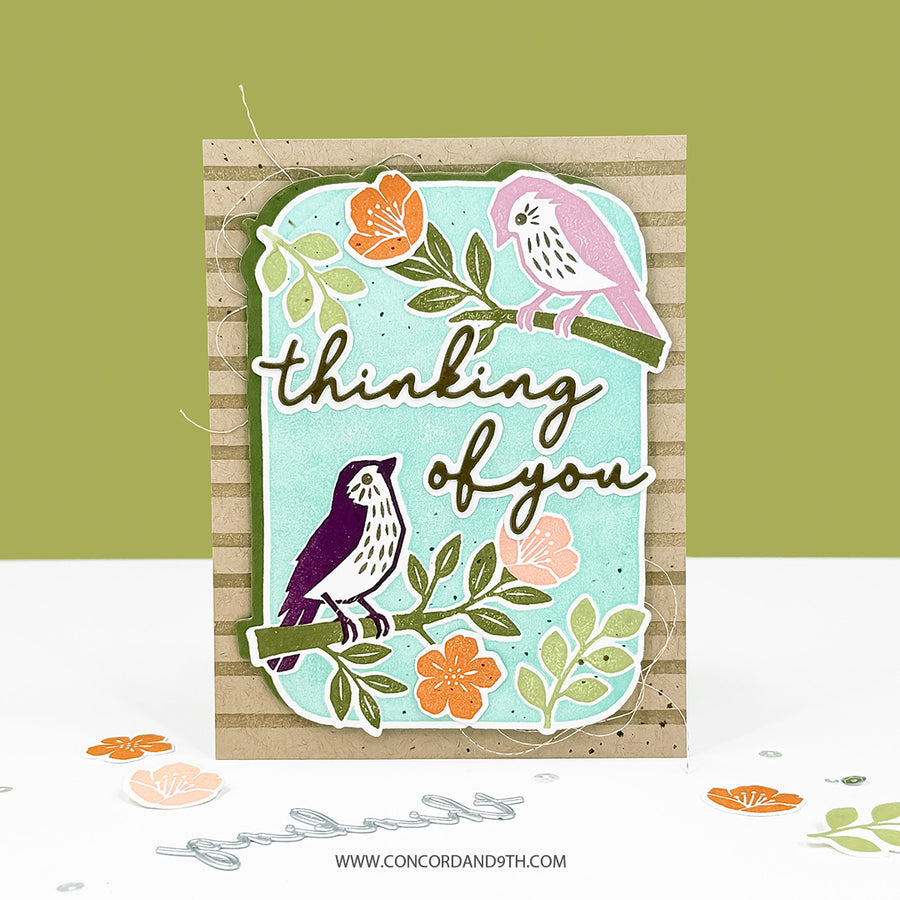 A handmade greeting card featuring two stylized birds perched on branches with the phrase 'thinking of you' in script. The card has a light aqua center panel layered over a tan striped backing, with orange and pink flowers and green leaves.