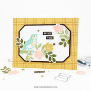 A handmade greeting card with a yellow plaid border, featuring a layered white central panel with a turquoise bird, pink and yellow flowers, green leaves, and the words 'MISS YOU' in small black blocks. The card is displayed on a white surface with crafting tools and paper pieces scattered around it.