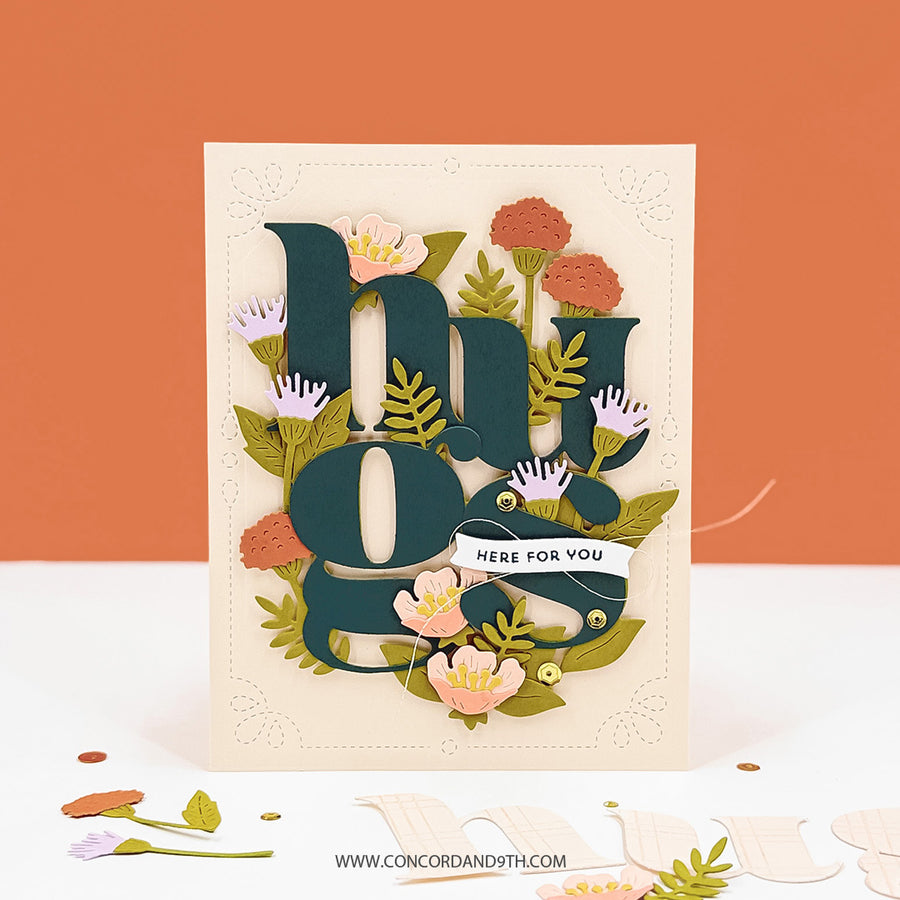 A handmade layered greeting card featuring the phrase 'hey guys' in dark teal letters surrounded by layered paper flowers and green leaves, with a small banner reading 'HERE FOR YOU'. The card is displayed on a white surface with scattered paper pieces against a solid orange background.