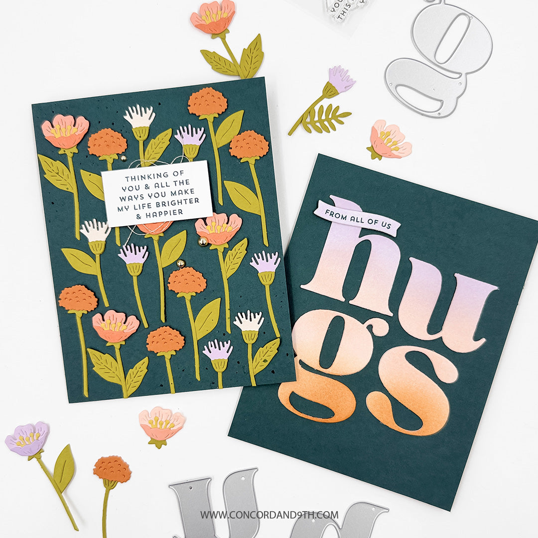 Two handmade greeting cards displayed on a white background with decorative paper flowers and metal die cutouts. One card features a dark teal background with illustrated orange, pink, and white flowers, a small white label reading 'THINKING OF YOU & ALL THE WAYS YOU MAKE MY LIFE BRIGHTER & HOPPIER,' and the word 'hugs' in large gradient letters on the other card.
