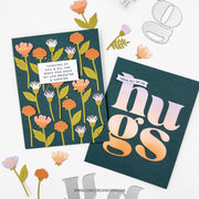 Two handmade greeting cards displayed on a white background with decorative paper flowers and metal die cutouts. One card features a dark teal background with illustrated orange, pink, and white flowers, a small white label reading 'THINKING OF YOU & ALL THE WAYS YOU MAKE MY LIFE BRIGHTER & HOPPIER,' and the word 'hugs' in large gradient letters on the other card.