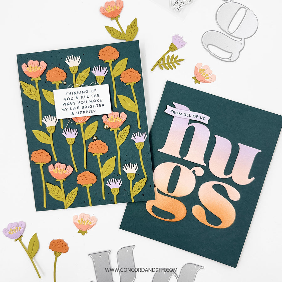 Two handmade greeting cards displayed on a white background with decorative paper flowers and metal die cutouts. One card features a dark teal background with illustrated orange, pink, and white flowers, a small white label reading 'THINKING OF YOU & ALL THE WAYS YOU MAKE MY LIFE BRIGHTER & HOPPIER,' and the word 'hugs' in large gradient letters on the other card.