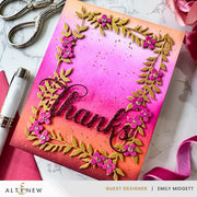 A handmade greeting card with a vibrant pink, orange, and yellow gradient background, featuring a raised metallic gold leaf floral border with pink flowers and the word 'thanks' in black script. The card is styled on a white marble surface with crafting tools like scissors, a pen, and ribbon.