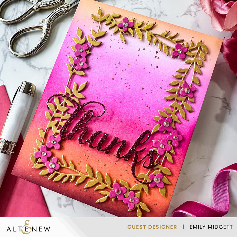 A handmade greeting card with a vibrant pink, orange, and yellow gradient background, featuring a raised metallic gold leaf floral border with pink flowers and the word 'thanks' in black script. The card is styled on a white marble surface with crafting tools like scissors, a pen, and ribbon.