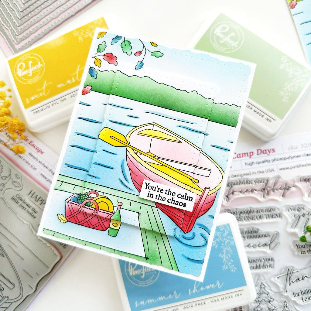 A colorful handmade greeting card featuring a pink and yellow rowboat on a blue water background, surrounded by other crafting supplies including ink pads, stamps, and clear sheets on a white surface.