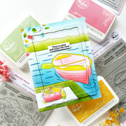 A handmade layered card featuring a pink boat on blue water with a yellow paddle, surrounded by Ranger brand ink pads and stamp designs on a white background.