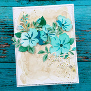 A handmade layered paper card featuring turquoise and teal flowers, green leaves, a beige watercolor wash background, and the script text 'Just a note' in gold foil, displayed on a distressed teal wooden surface.