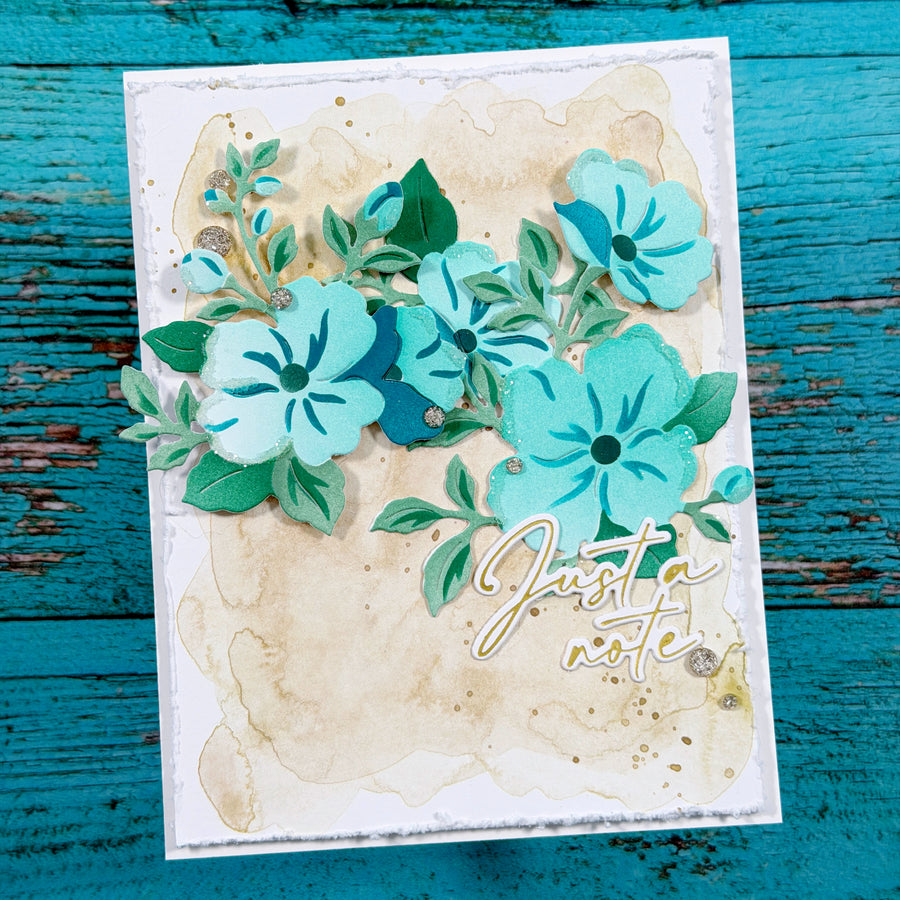 A handmade layered paper card featuring turquoise and teal flowers, green leaves, a beige watercolor wash background, and the script text 'Just a note' in gold foil, displayed on a distressed teal wooden surface.