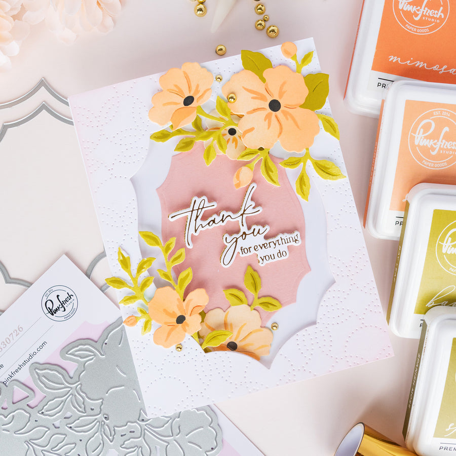 A handmade layered card with peach and yellow flowers, green leaves, and a pink center panel reading 'thank you for everything you do,' styled with crafting tools and ink pads on a pale pink background.