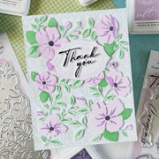 A handmade layered card featuring purple and green floral designs with a central white label reading 'Thank you' in black script. The card is styled on a desk with crafting tools and stamps visible around it.