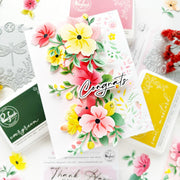 A beautifully arranged flat-lay of handmade floral greeting cards and crafting supplies, featuring a central white card with layered 3D flowers in pink, yellow, and green, and the word 'Congrats' in white script. Surrounding items include ink pads, metal dies, and other cards on a white background.