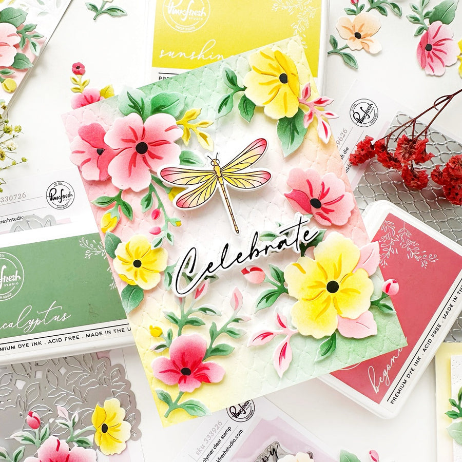 A handmade floral card with layered pink and yellow flowers, green leaves, a dragonfly embellishment, and the word 'Celebrate' in script, styled on top of other craft stamp and ink boxes from Handmade in Philadelphia.