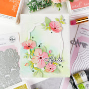A handmade greeting card with a pastel green and white watercolor wash background, featuring layered white scalloped frames, pink flowers, green leaves, and the word 'Hello' in black script. The card is displayed in a flat-lay arrangement of crafting supplies, including Ranger Studio stamping and die sets, and other craft products.