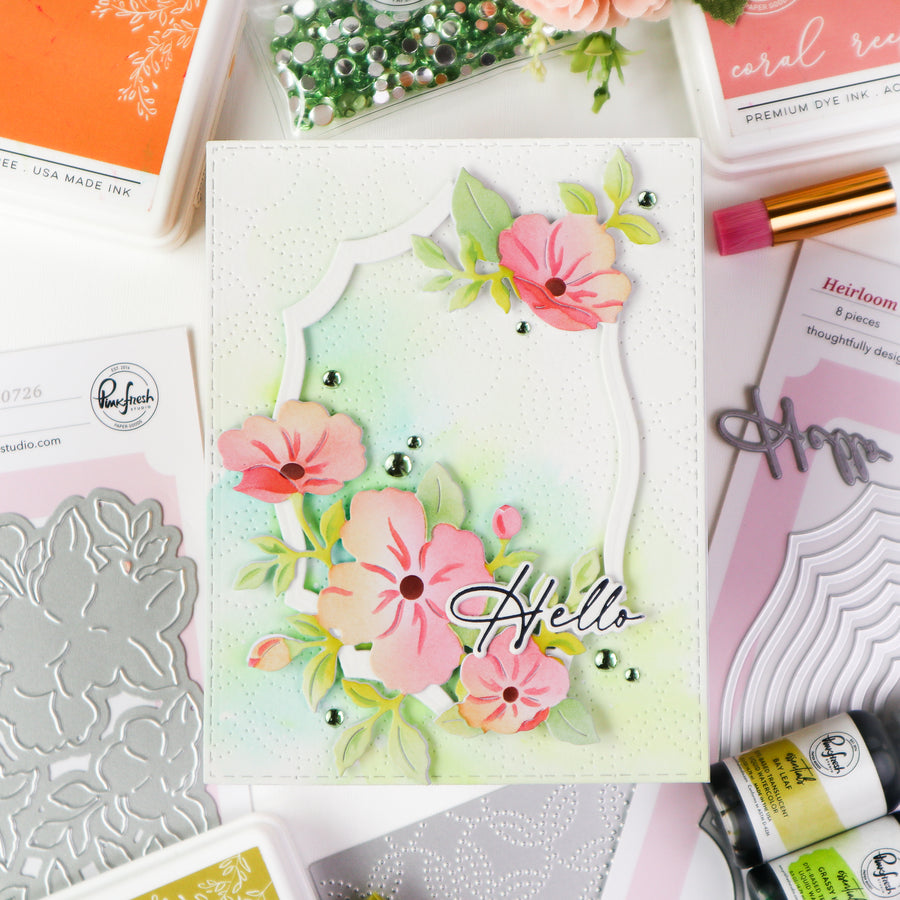 A handmade greeting card with a pastel green and white watercolor wash background, featuring layered white scalloped frames, pink flowers, green leaves, and the word 'Hello' in black script. The card is displayed in a flat-lay arrangement of crafting supplies, including Ranger Studio stamping and die sets, and other craft products.