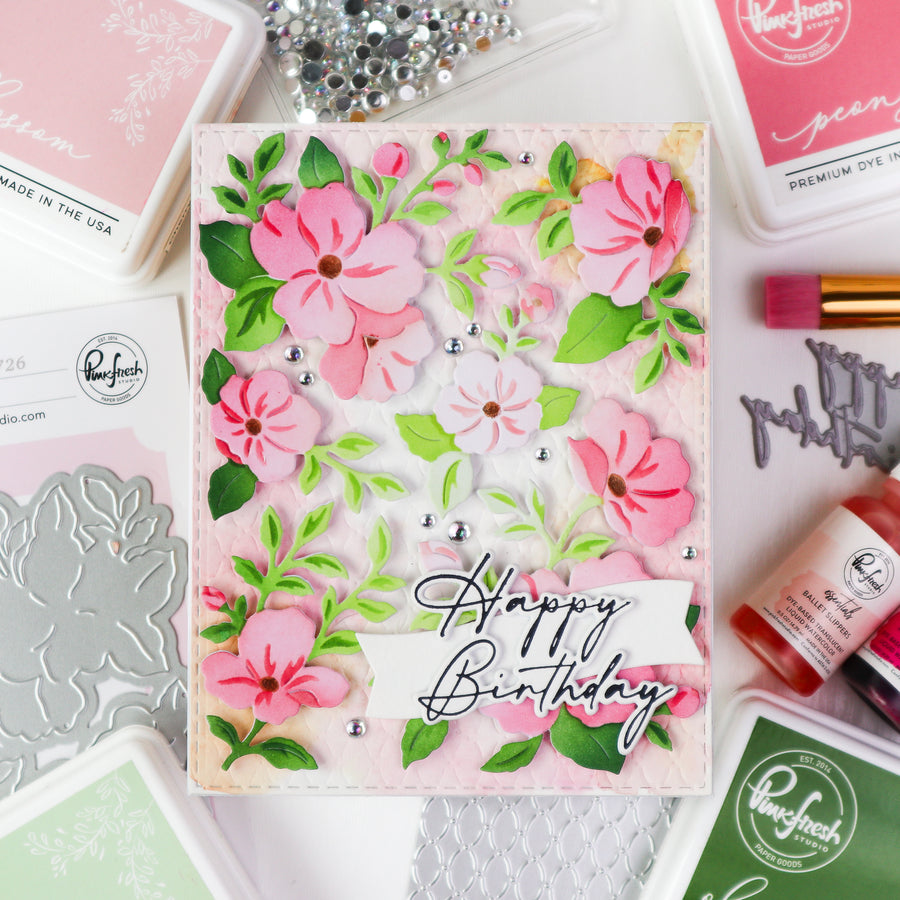 A handmade layered card with pink flowers and green leaves on a textured light pink background, featuring a white banner that reads 'Happy Birthday' in black script. The card is styled in a flat-lay with crafting supplies including Ranger ink pads, rhinestones, and metal dies.