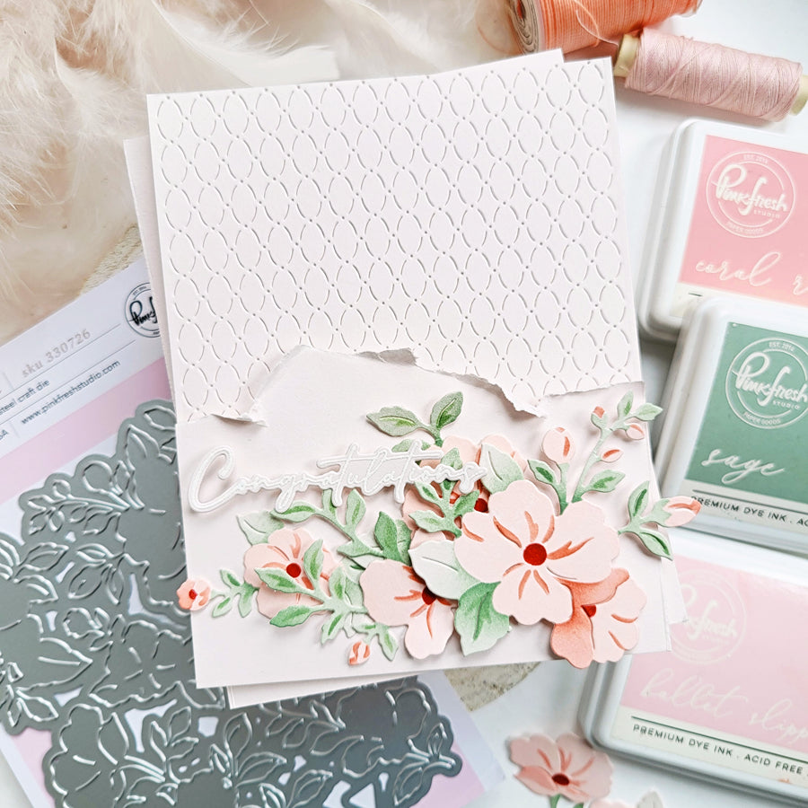 A handmade layered card featuring a white scalloped embossed flap, a white cursive 'Congratulations' script, and 3D floral die-cut elements in coral pink, peach, and sage green. The card is styled on a white surface with craft supplies, including a silver metal die, pink and green Ranger ink pads, and peach thread spools.
