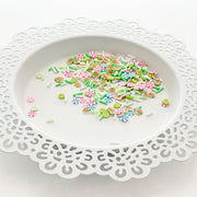 A pile of colorful craft embellishments, including flower charms, sprinkles, and small decorative pieces, displayed on a white scalloped plate against a white background. Pretty Pink Posh Gingerbread Cheer Clay Confetti