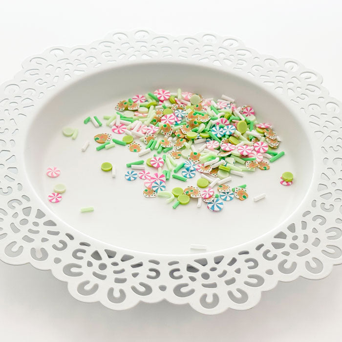 A pile of colorful craft embellishments, including flower charms, sprinkles, and small decorative pieces, displayed on a white scalloped plate against a white background. Pretty Pink Posh Gingerbread Cheer Clay Confetti