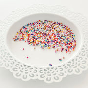 Multicolor craft sprinkles or craft beads scattered on a white ceramic plate with a scalloped edge, set against a clean white background. Pretty Pink Posh Gingerbread Lane Shaker Beads