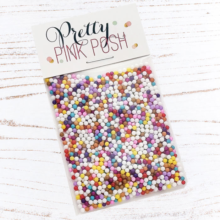 A clear plastic bag filled with many small round craft sprinkles in assorted pastel and bright colors, attached to a white header card that reads 'Pretty Pink Posh.' The package is photographed on a white wood surface. Pretty Pink Posh Gingerbread Lane Shaker Beads