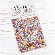 A clear plastic bag filled with many small round craft sprinkles in assorted pastel and bright colors, attached to a white header card that reads 'Pretty Pink Posh.' The package is photographed on a white wood surface. Pretty Pink Posh Gingerbread Lane Shaker Beads