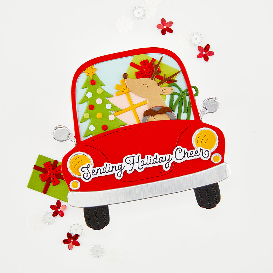 A festive red car handmade card with a reindeer driving it, holiday decorations inside, gift boxes, and the phrase 'Sending Holiday Cheer' on the front, set on a light gray background with scattered snowflakes and red flowers. 