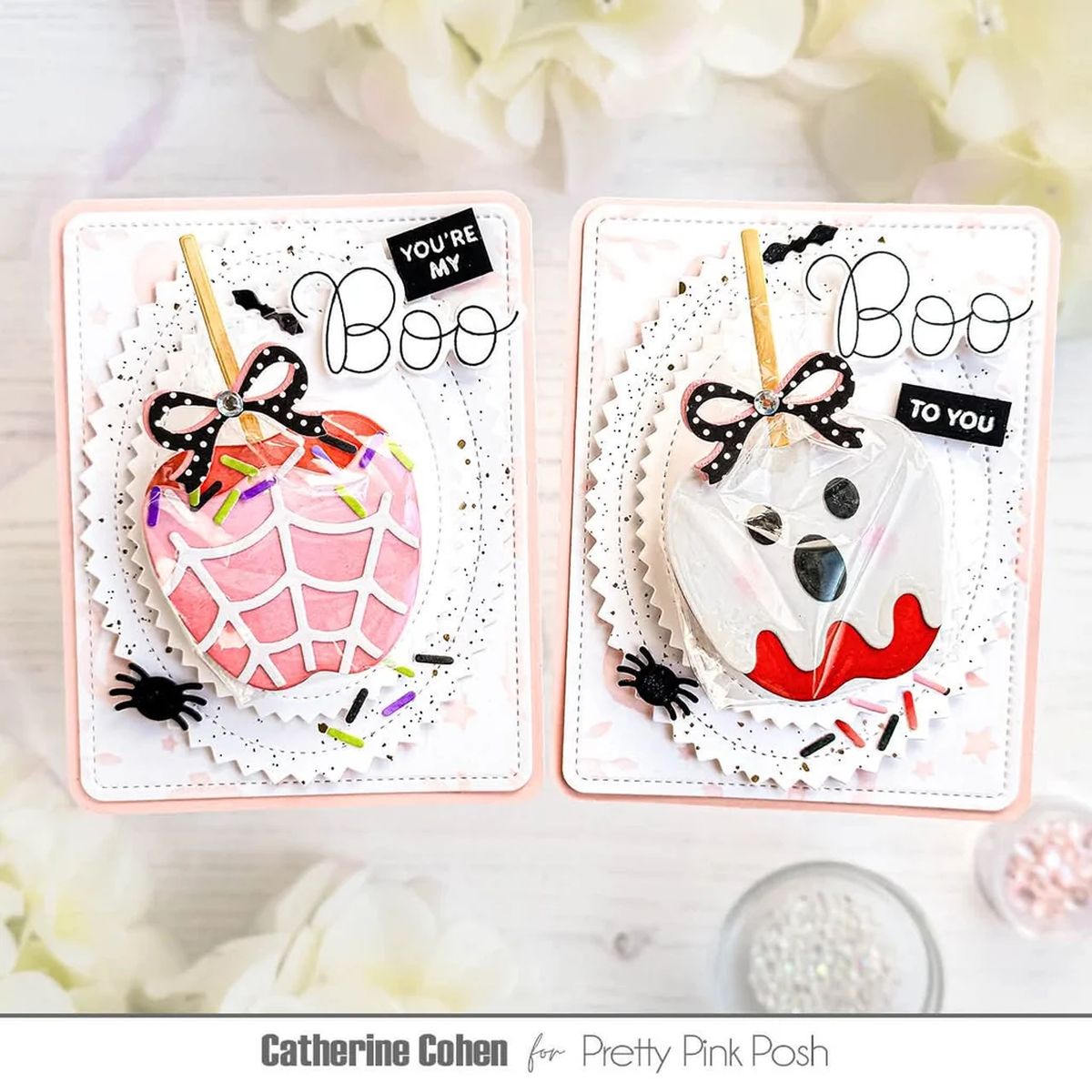 Two Halloween-themed greeting cards displayed side by side on a white surface with soft floral props. The left card shows a pink spiderweb-covered apple with black spider and bow details, and the right card shows a ghost face with black-and-white polka dots and red lips.