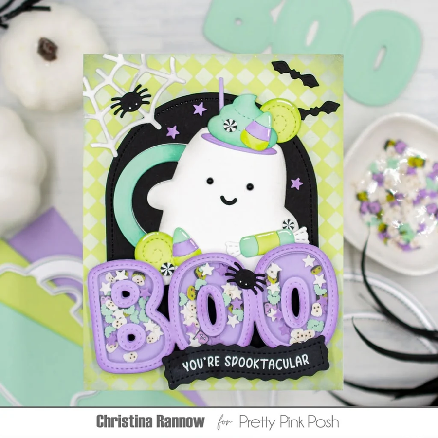 A handmade Halloween greeting card featuring a smiling white ghost wearing a green and purple witch hat, surrounded by black bats, purple 'BOO' letters, and a black banner reading 'YOU'RE SPOOKTACULAR'. The card is displayed on a styled flat-lay background with white pumpkins, pastel decorations, and a small dish of glitter.
