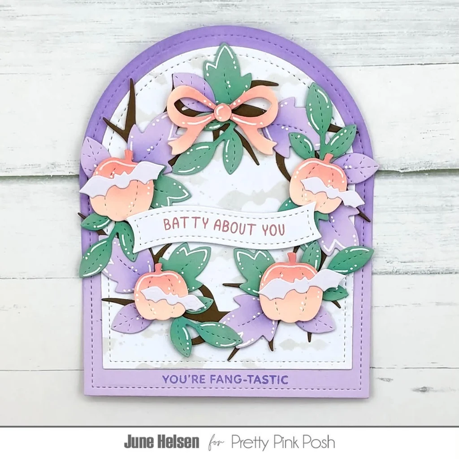A handmade layered card featuring a lavender arched border, a white center, and a circular wreath design made from purple paper flowers, green leaves, peach-colored peaches, and brown branches. A white banner across the wreath reads 'BATTY ABOUT YOU', and the bottom border reads 'YOU'RE FANG-TASTIC'.