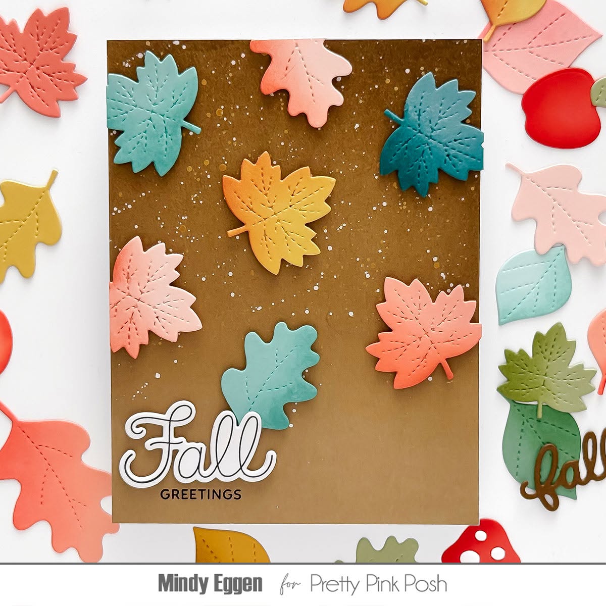 A handmade autumn-themed greeting card with colorful layered maple leaves on a brown background, surrounded by scattered matching leaves and decorative elements on a white surface. The card reads 'Fall Greetings' in white script and block text.
