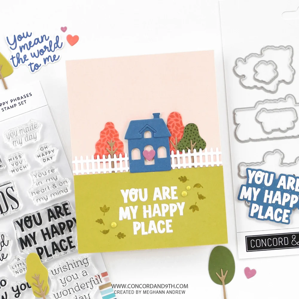 A handmade greeting card with a layered paper design of a blue house, white picket fence, and colorful trees on a yellow-green lawn, displayed alongside stamp sets, die cut metal shapes, and sticker elements on a white background.