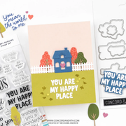 A handmade greeting card with a layered paper design of a blue house, white picket fence, and colorful trees on a yellow-green lawn, displayed alongside stamp sets, die cut metal shapes, and sticker elements on a white background.