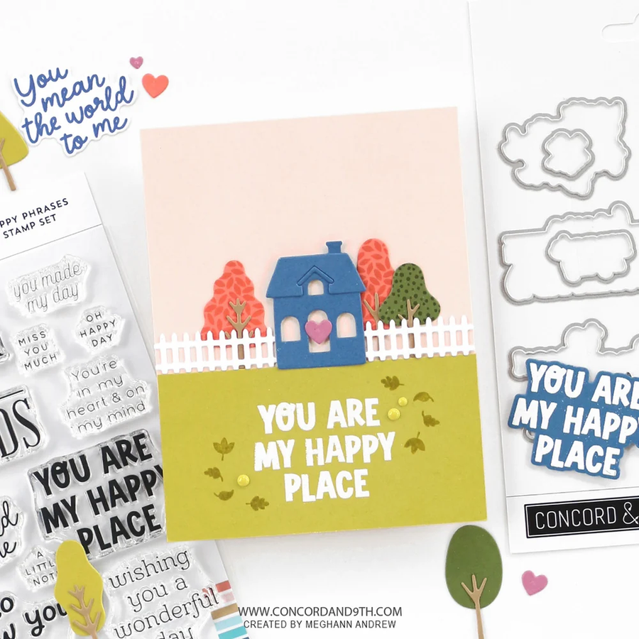 A handmade greeting card with a layered paper design of a blue house, white picket fence, and colorful trees on a yellow-green lawn, displayed alongside stamp sets, die cut metal shapes, and sticker elements on a white background.