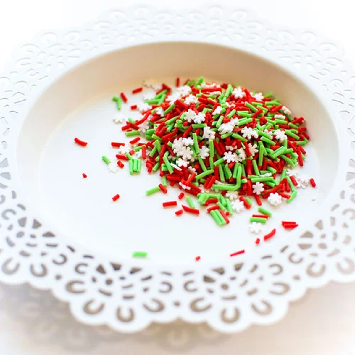 A pile of red, green, and white holiday sprinkles, including star and snowflake shapes, resting on a white ceramic plate with a scalloped cutout edge. Product is called Holiday Cheer Clay Confetti
