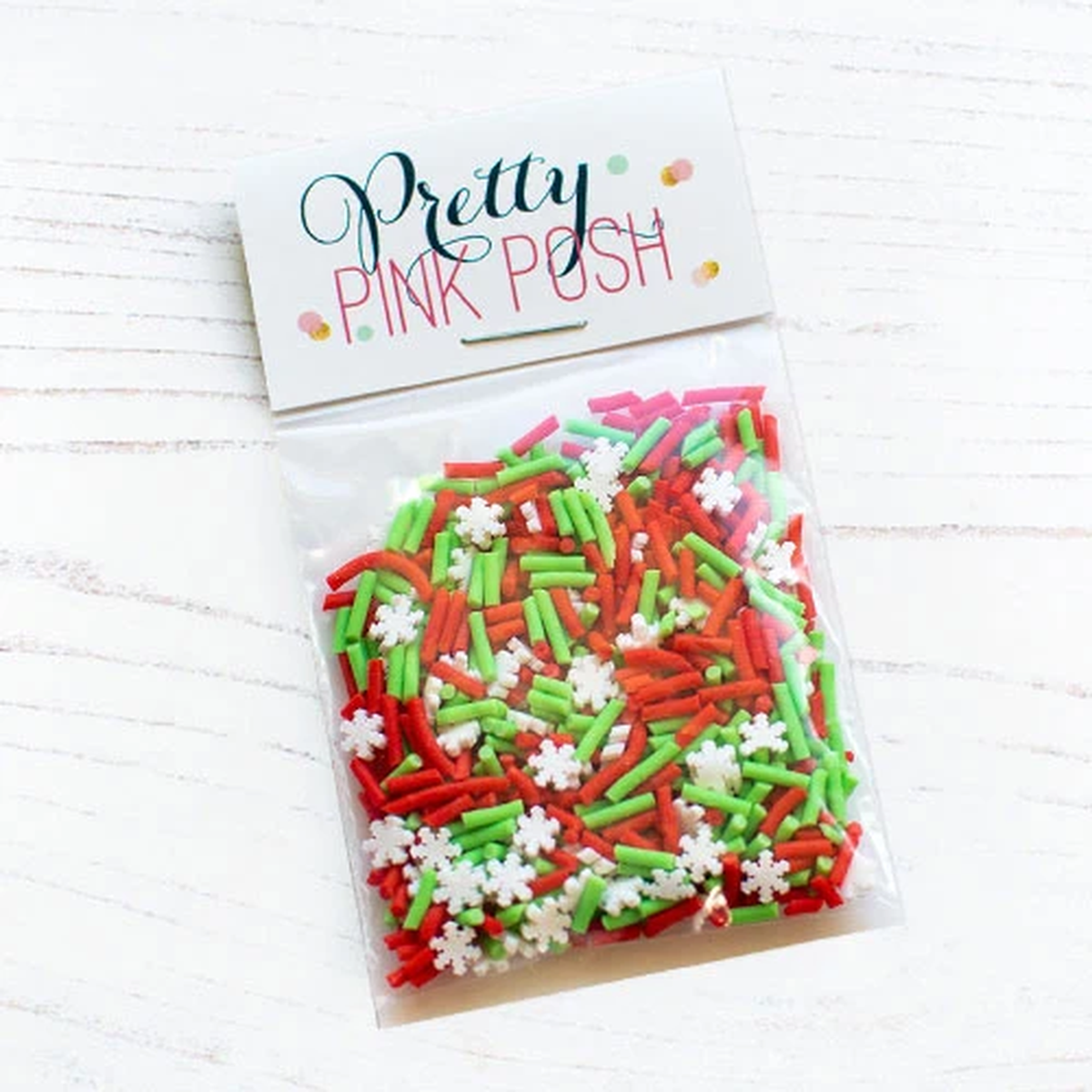 A clear plastic bag filled with red, green, and white sprinkles, sealed with a white paper header card that reads 'Pretty Pink Posh' on a light wood background. Product is called Holiday Cheer Clay Confetti