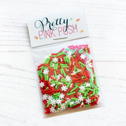 A clear plastic bag filled with red, green, and white sprinkles, sealed with a white paper header card that reads 'Pretty Pink Posh' on a light wood background. Product is called Holiday Cheer Clay Confetti