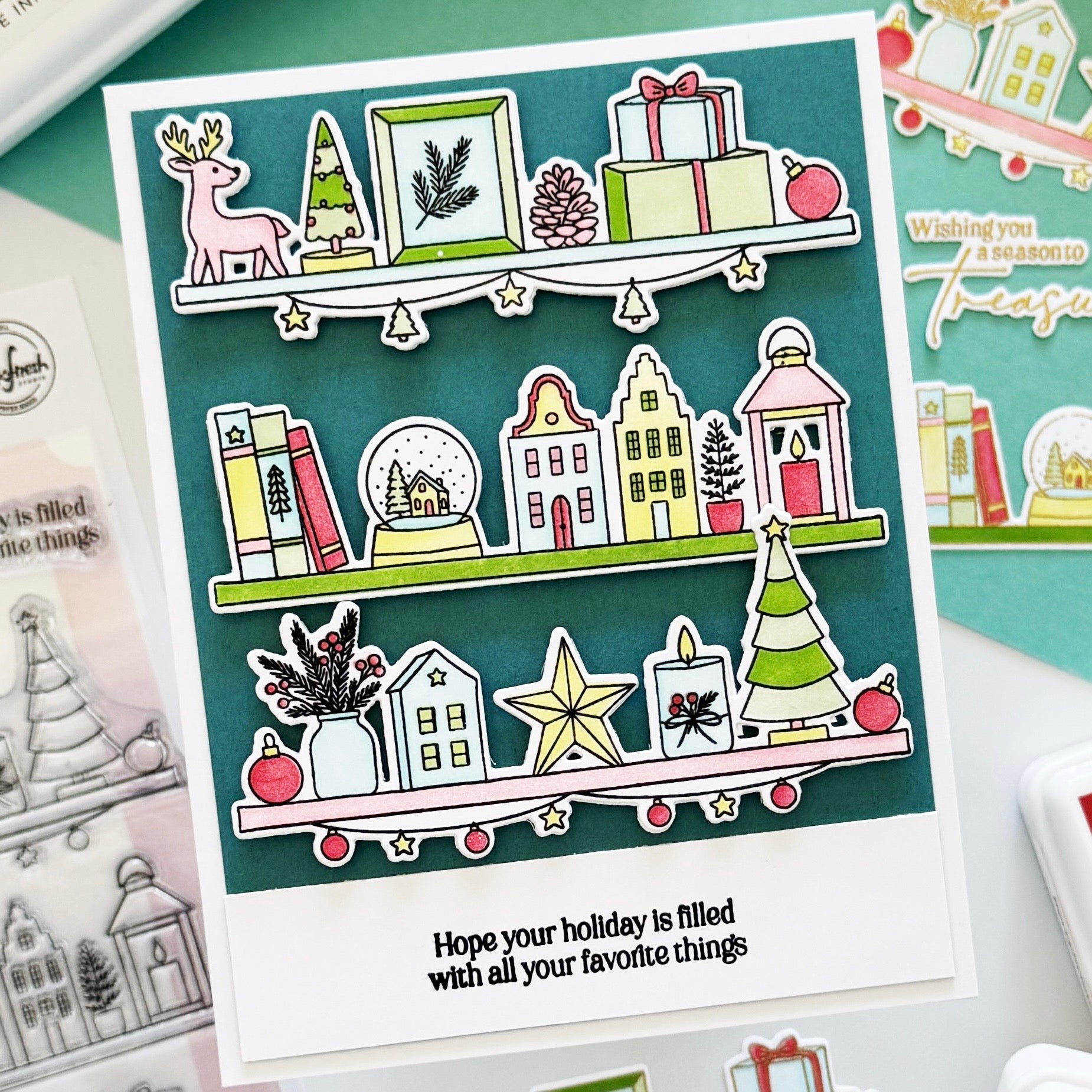 A handmade holiday greeting card featuring illustrated Christmas shelves with a deer, trees, gifts, houses, books, and ornaments, plus the printed phrase 'Hope your holiday is filled with all your favorite things.' The card is photographed on a styled flat-lay with other cards and stamp-related items around it.