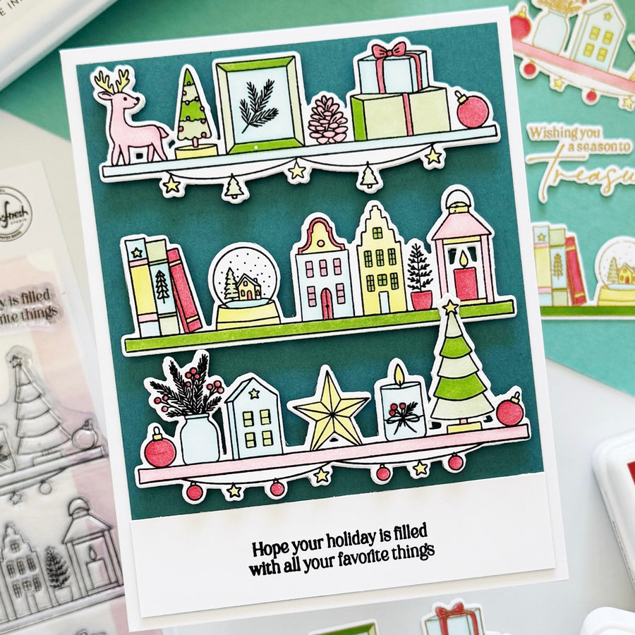 A handmade holiday greeting card featuring illustrated Christmas shelves with a deer, trees, gifts, houses, books, and ornaments, plus the printed phrase 'Hope your holiday is filled with all your favorite things.' The card is photographed on a styled flat-lay with other cards and stamp-related items around it.