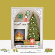 A 3D layered papercraft holiday card set featuring a fireplace scene with a Christmas tree, gift boxes, a burning log, and a 'Happy Holidays' banner. The card is displayed on a white surface against a mustard yellow background, with a few small craft pieces scattered nearby.
