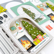 A close-up of a handmade layered paper craft featuring a Christmas tree, fireplace, and holiday gifts, displayed alongside other scrapbooking and card-making supplies on a white surface.