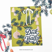 A handmade Christmas greeting card with a green background, layered pine branches, hanging beige bells, and the phrase 'Deck the Halls' in layered white and navy lettering. The card is styled on a white background with holiday paper decorations around it.